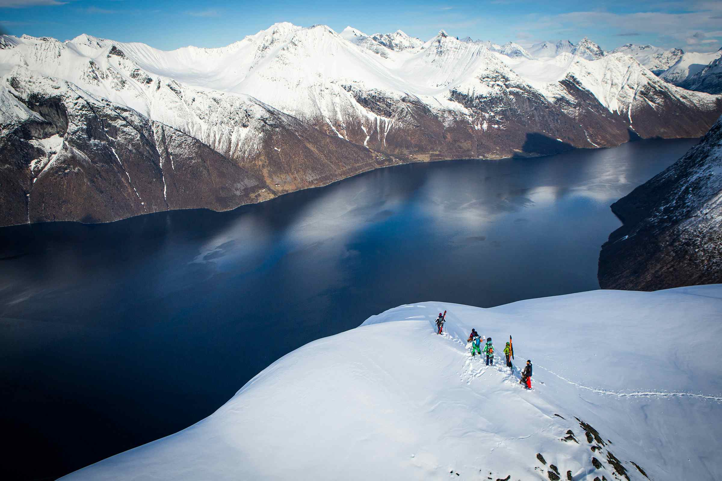 Personer på topptur med ski på fjelltopp med utsikt over blå fjord.