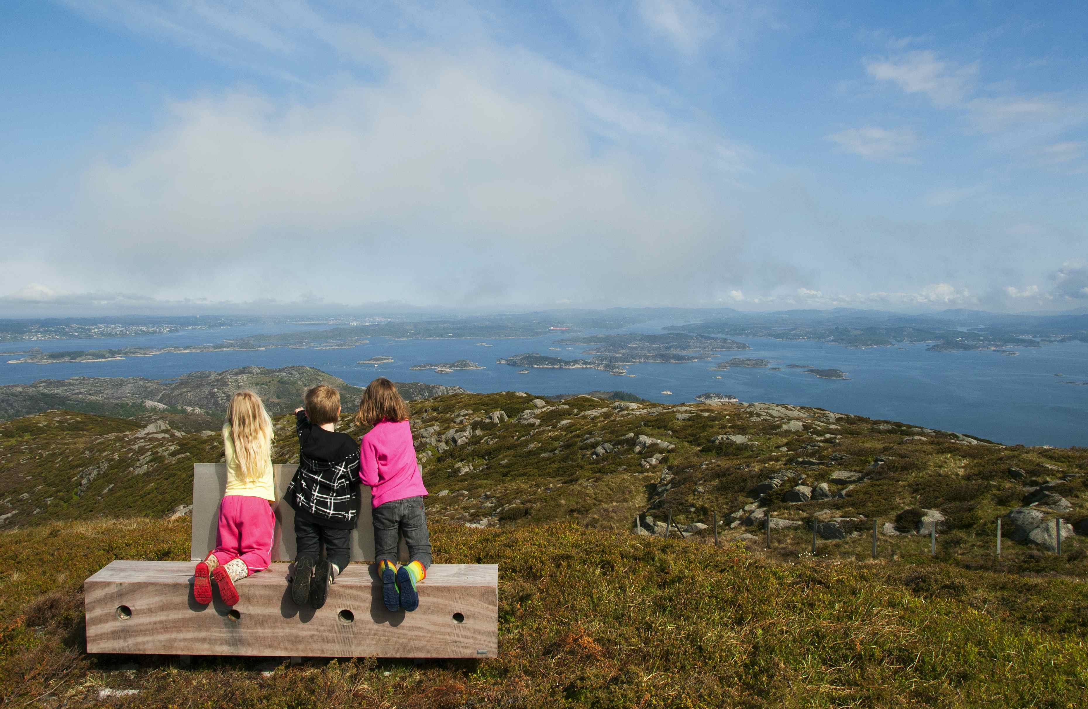 Tre barn som sitter på knærne på en benk med ryggen til, og ser på utsikten over hav og øyer.