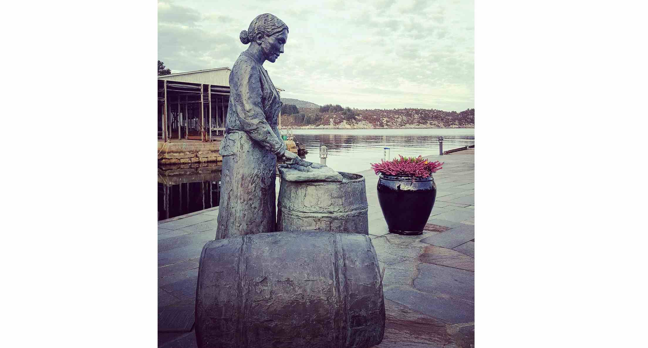Statue av jente som rensker sild på tønnefat ved sjø.