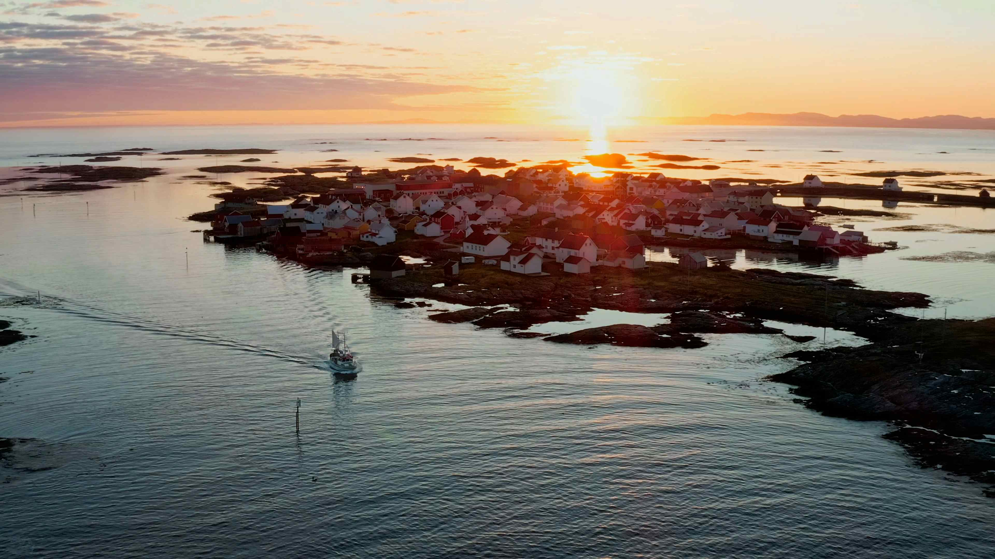 Fiskeværet Veiholmen med havet og solnedgangen i bakgrunnen, og en fiskebåt på vei inn i forgrunnen.