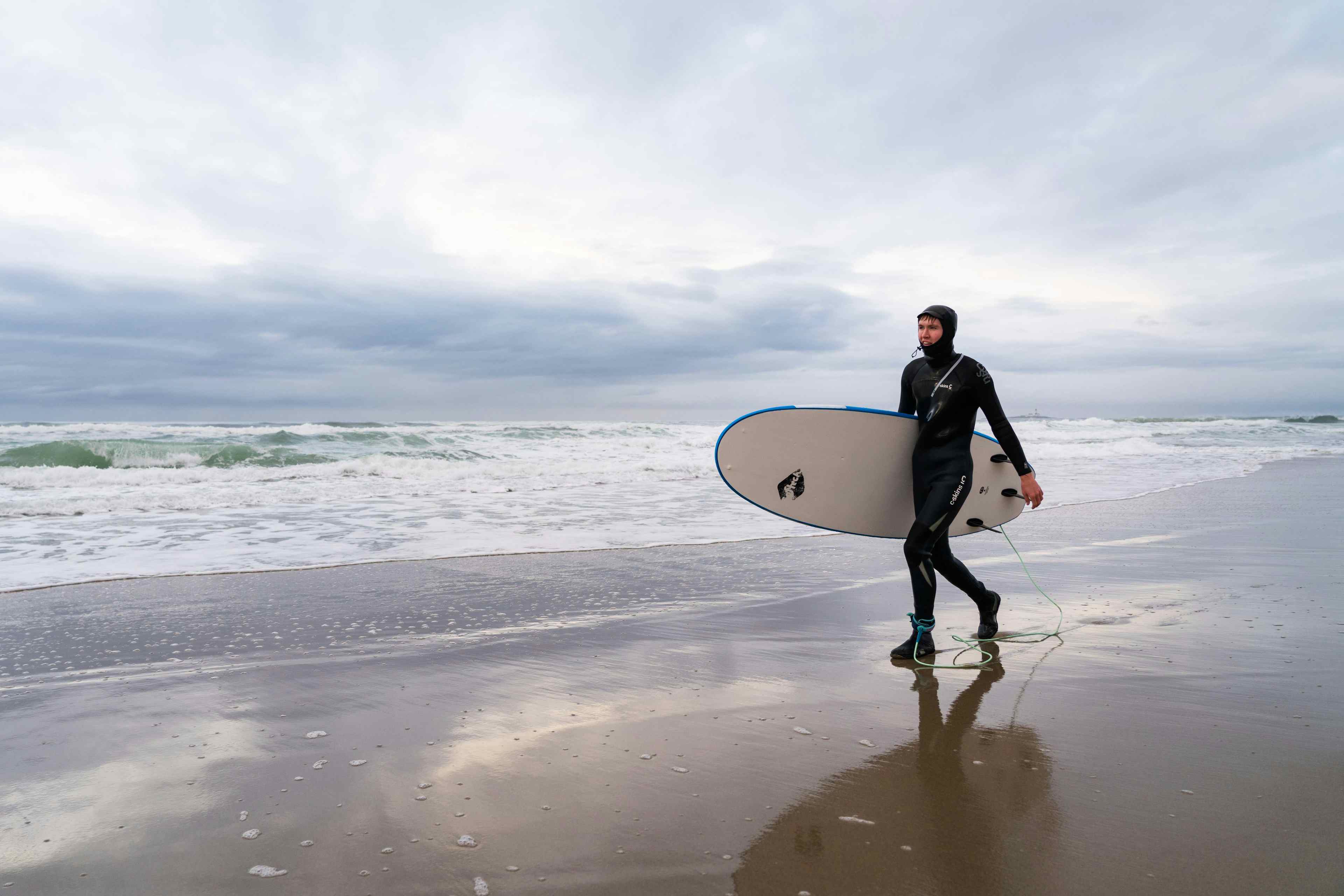 Mann i våtdrakt går langs stranden med surfebrett under armen.