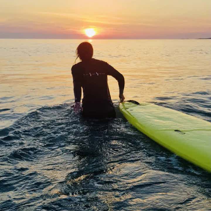 Dame i solnedgang med surfebrett på vei ut i sjøen.