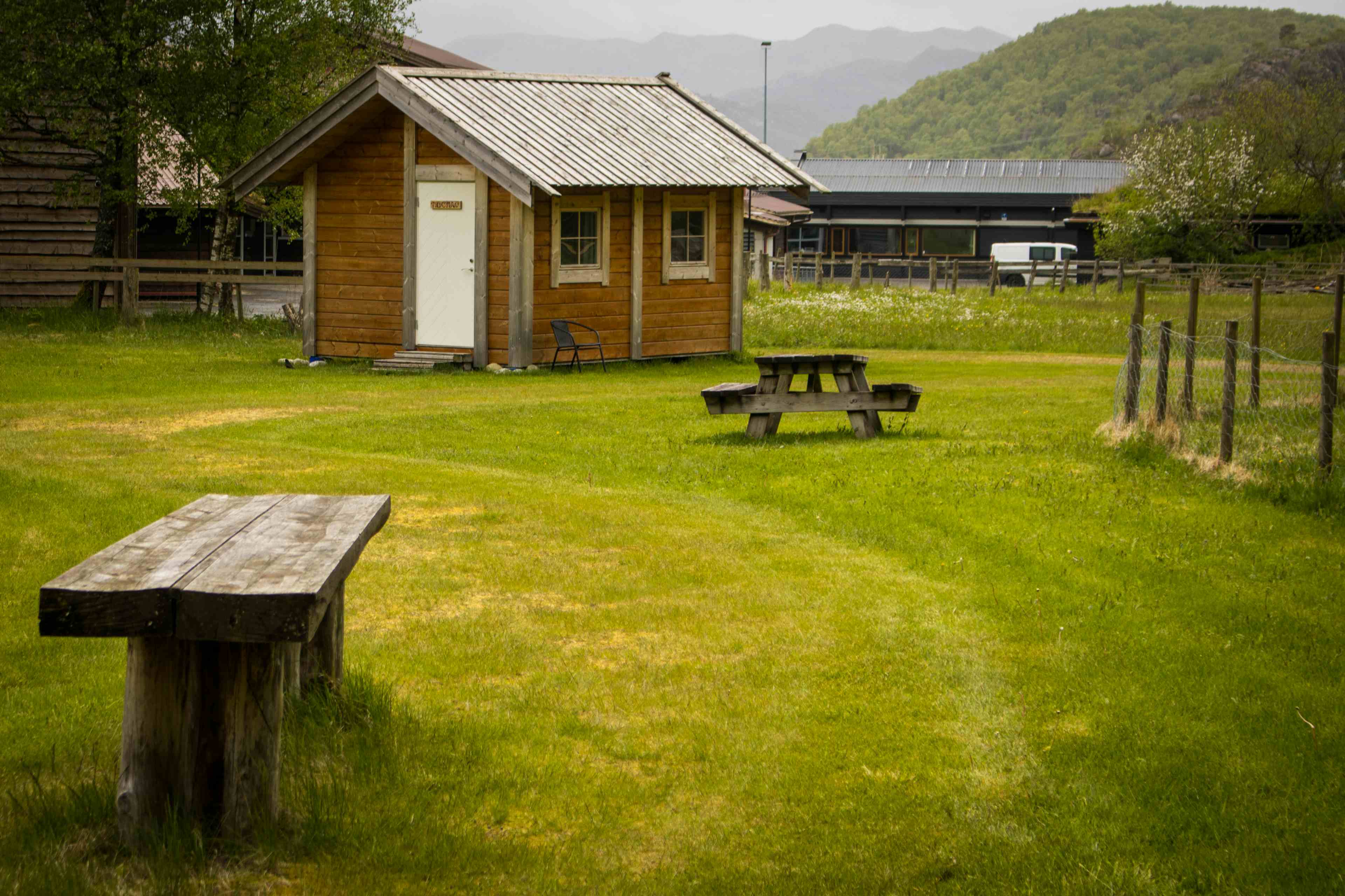 To hytter og sanitæranlegget i bakgrunnen og et grønt område med benker og bord i forgrunnen.