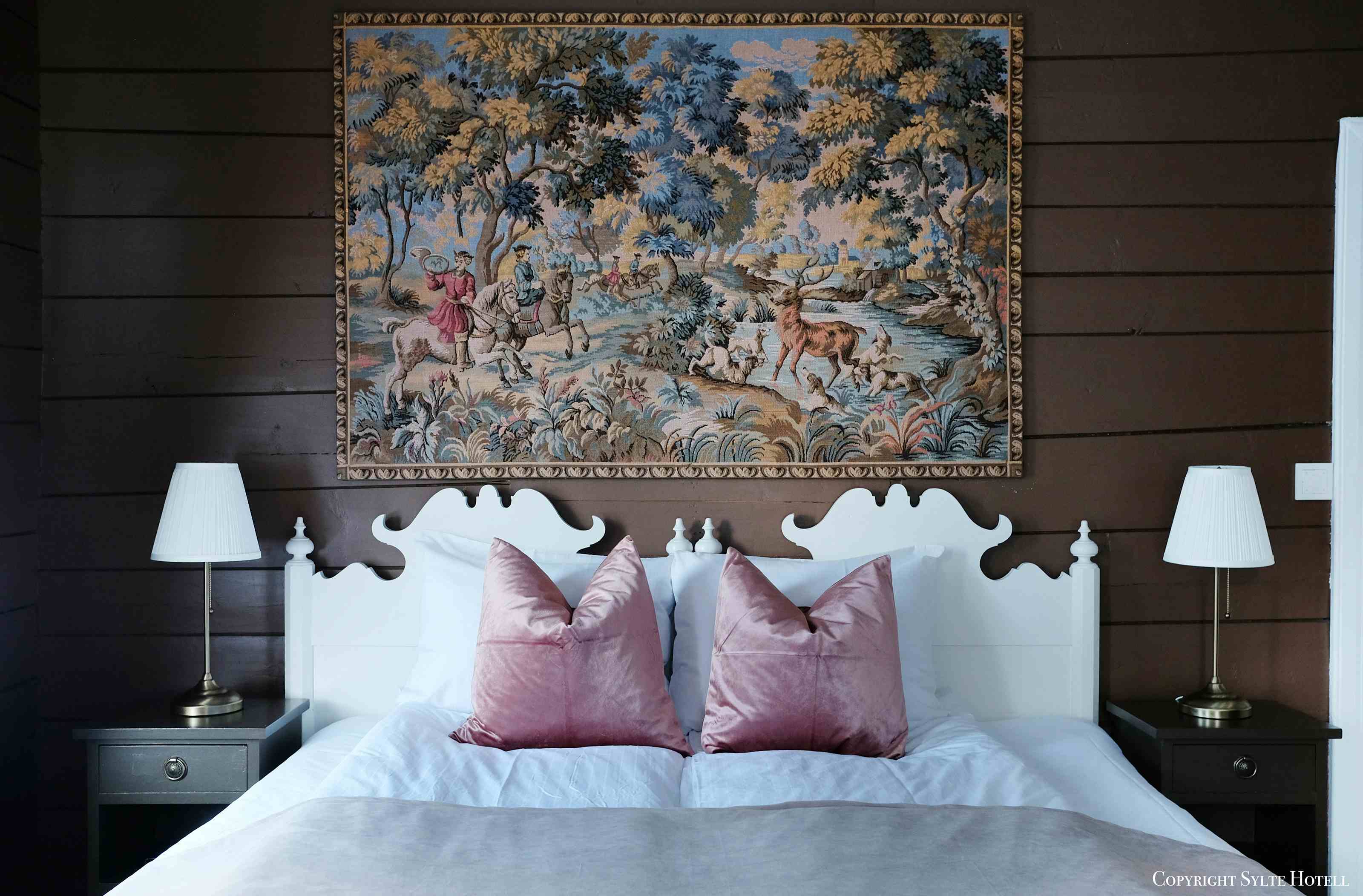 En seng i toner av rosa, med et stort broderi over sengen med historisk motiv.