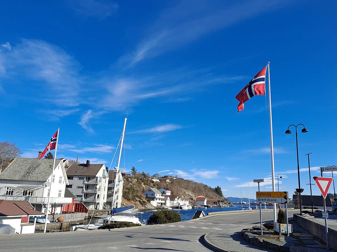 Blå sjø, asfaltert veg rundt sjøen, hvite hus, vegskilt og to flaggstenger med norske flagg på.