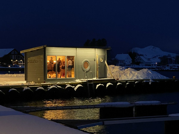 A sauna located at a pier - seen from a distance with people inside. It's evening on the photo.