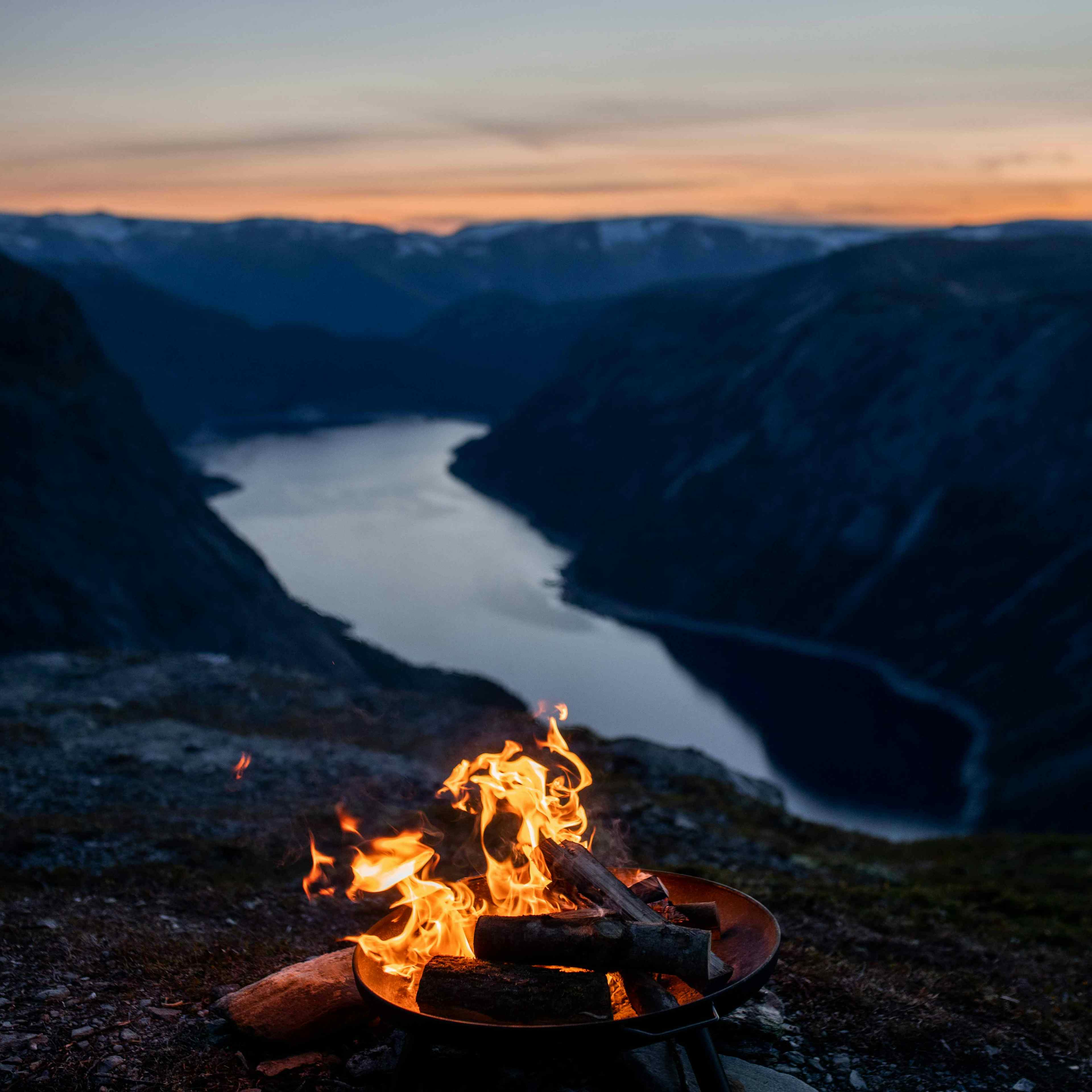 Bålpanne med åpen ild på en fjelltopp ved solnedgang med utsikt over en fjord.