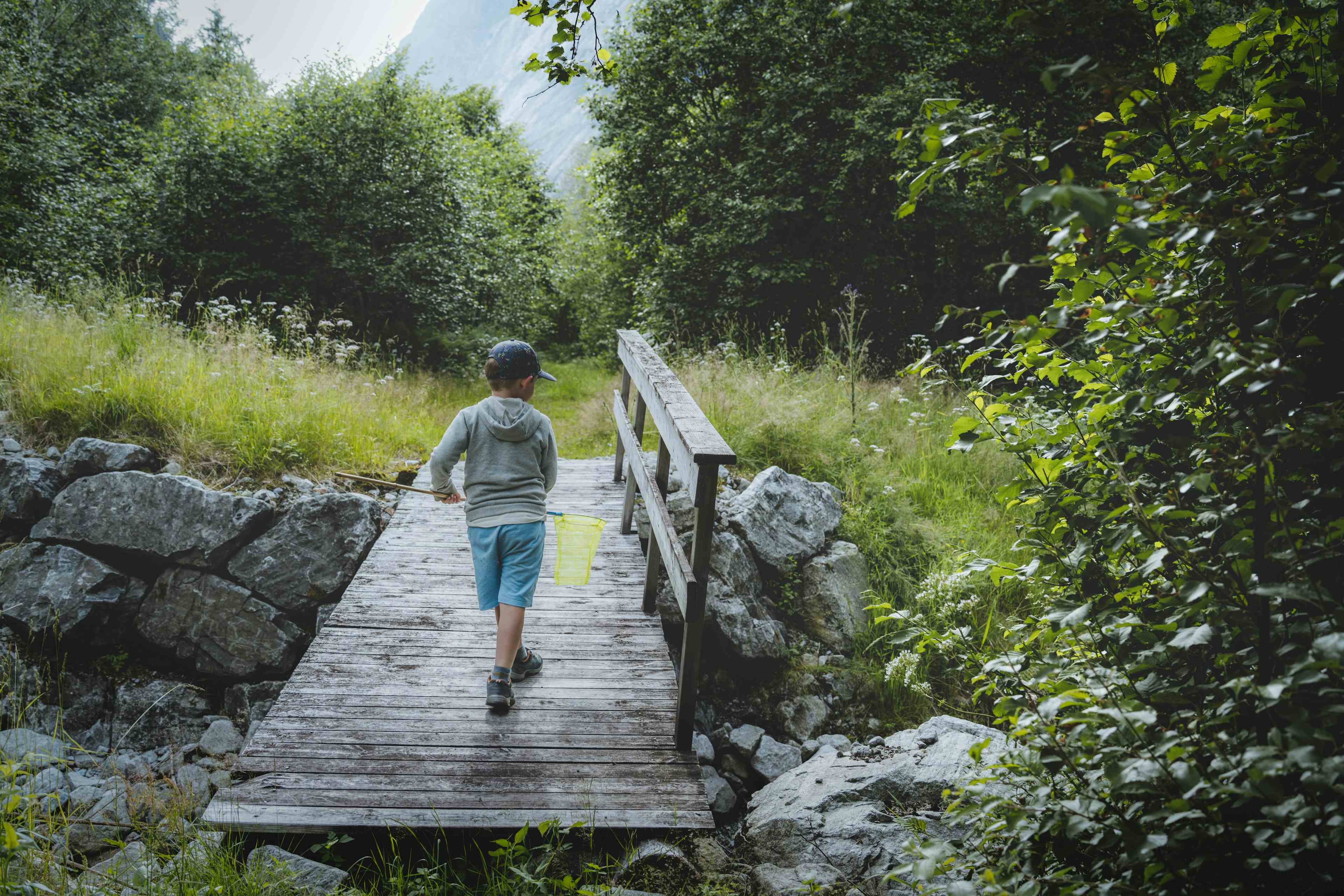 En liten gutt med en hov går over en bro i skogen.
