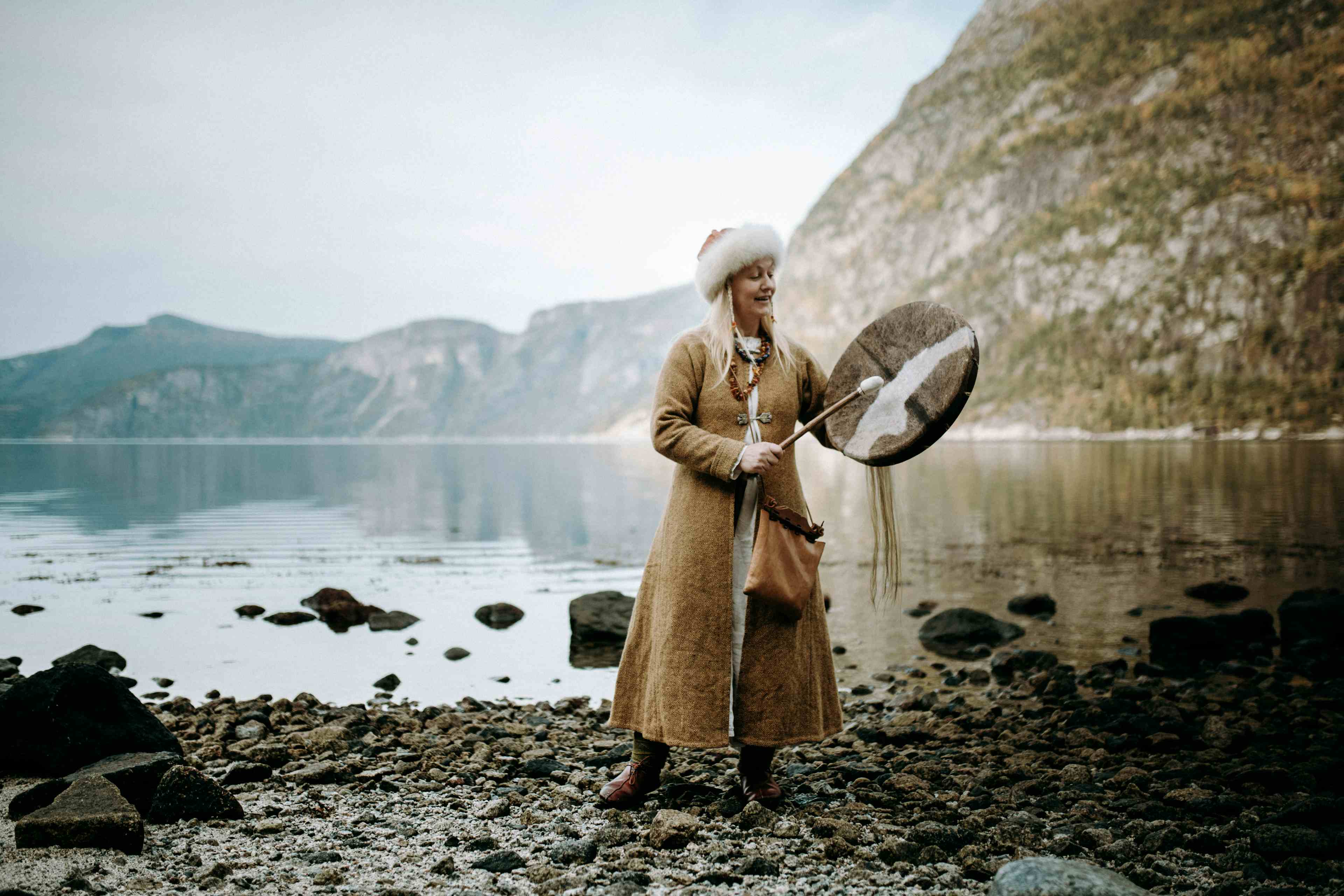 En kvinne utkledd som en viking, som står ved fjorden og spiller en vikingtromme.
