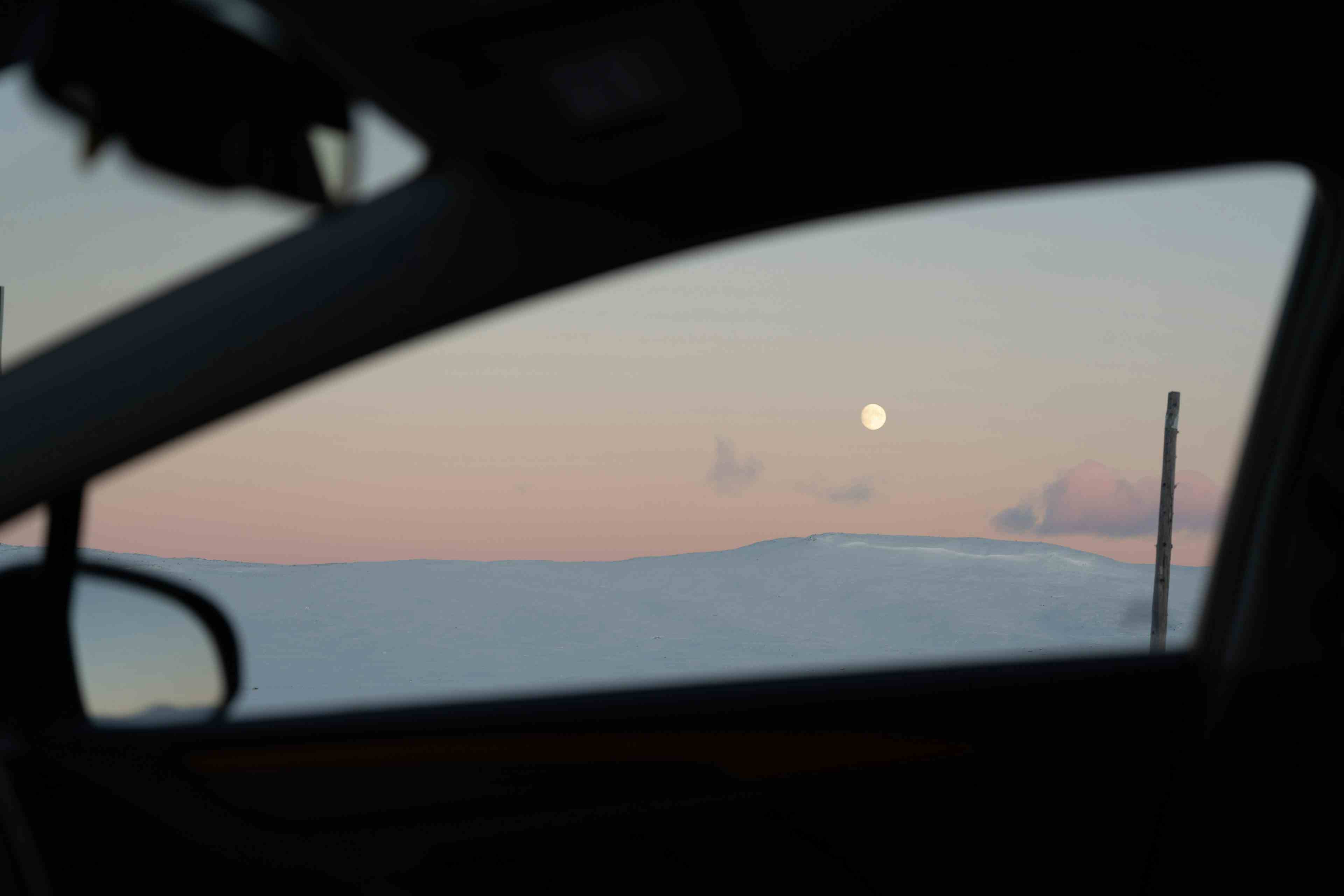 En bil kjører i snølandskapet over Hardangervidda, under en knallrosa himmel under fullmåne.