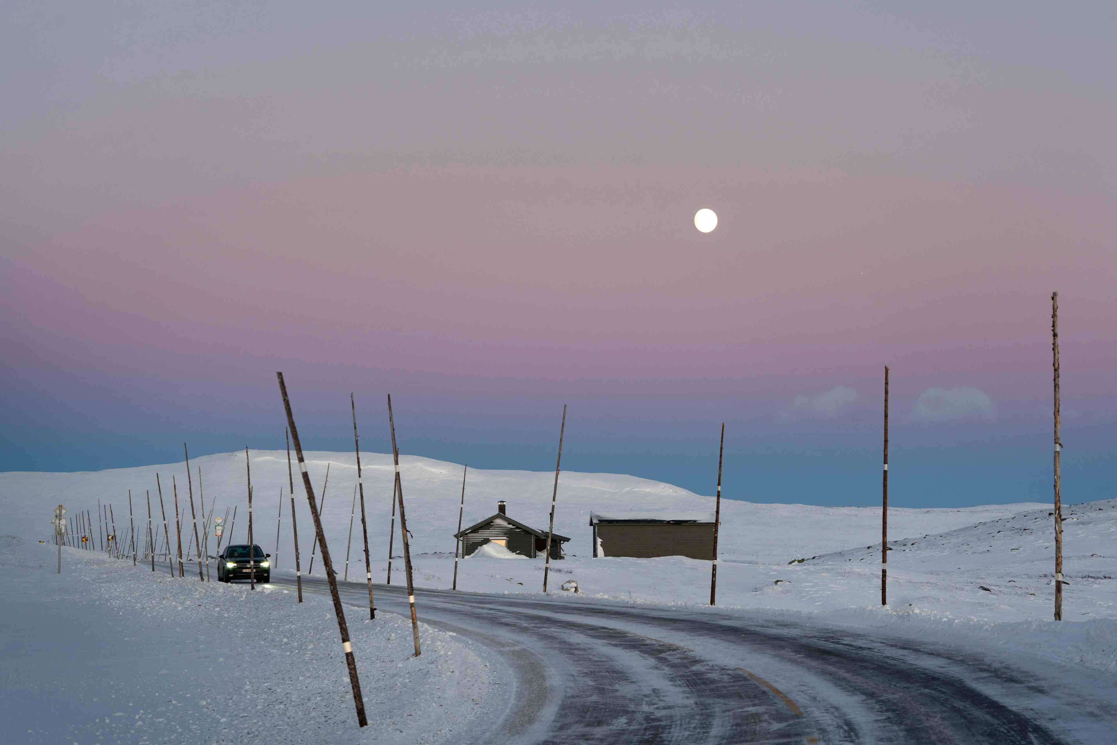 En bil kjører i snølandskapet over Hardangervidda, under en knallrosa himmel under fullmåne.
