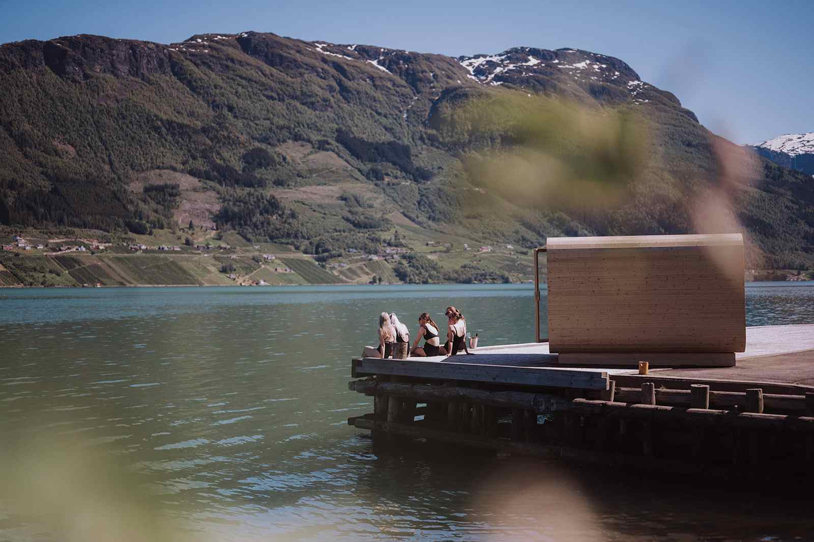 Personer nyter fjordbad på brygge foran Heit Grimo sauna med fjell i bakgrunnen
