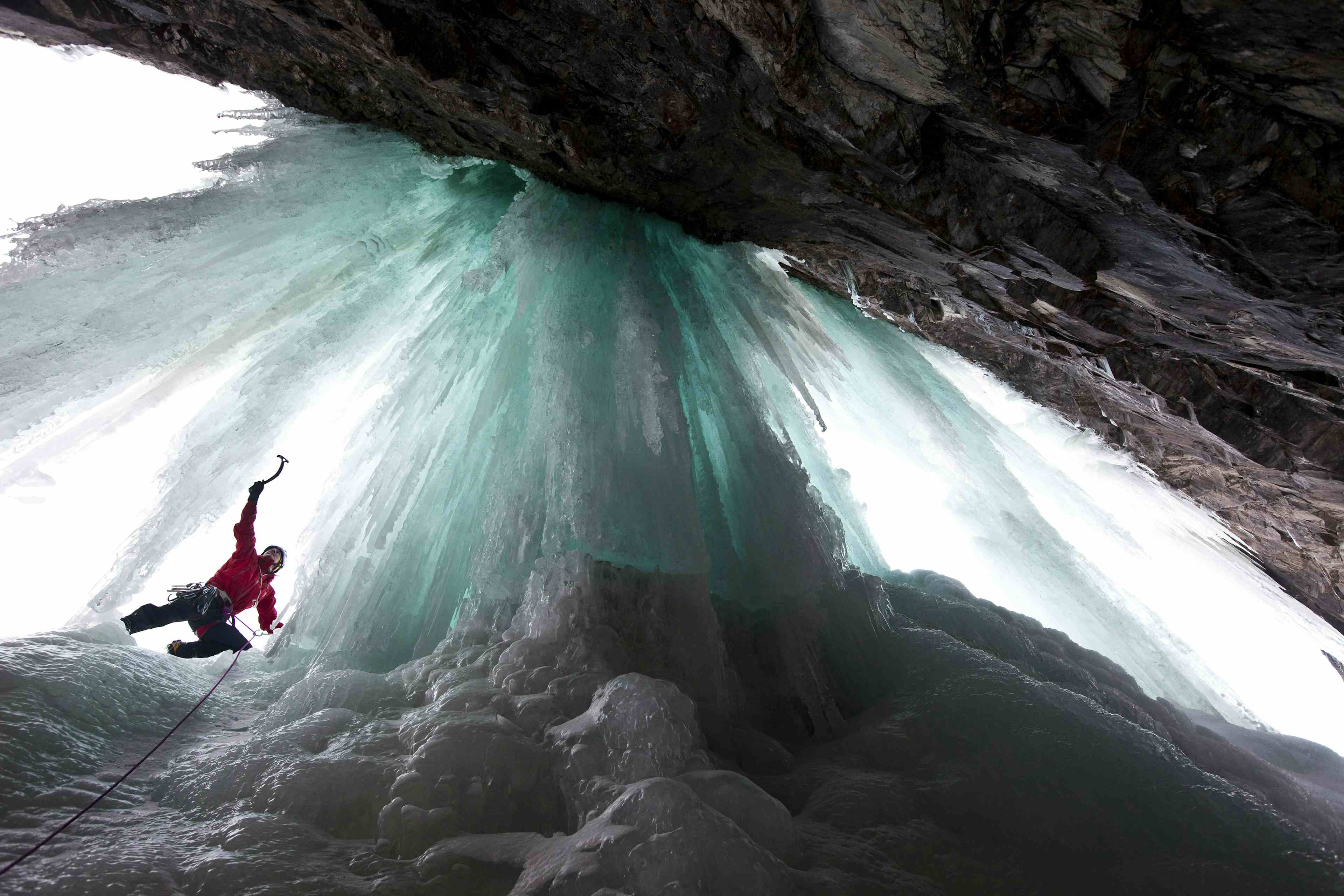 En isklatrer i rød jakke sett nedenfra klatrer den turkisfargede isen.